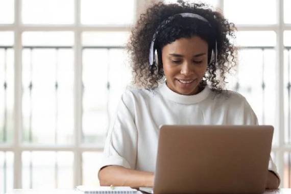 A black woman with headphones and a white jumper sitting at a desk and smiling as she types on her laptop