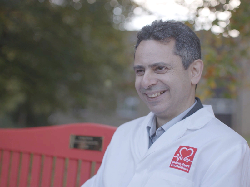 Professor Charalambos Antoniades sitting on a red bench smiling