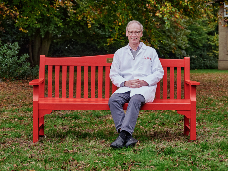 Professor Hugh Watkins sitting on a red bench smiling