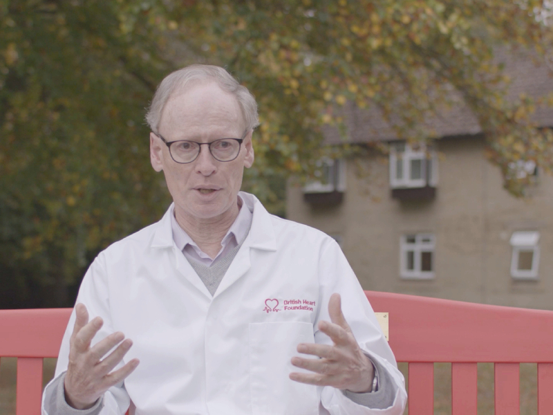 Professor Hugh Watkins sitting on a red bench discussing his research