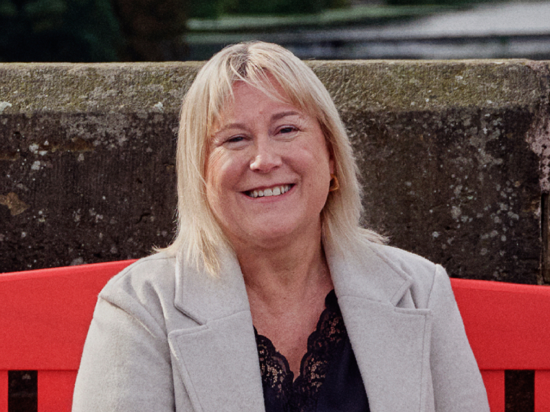 Louise is smiling while sitting on a red bench for BHF's Keep Us Beating campaign.