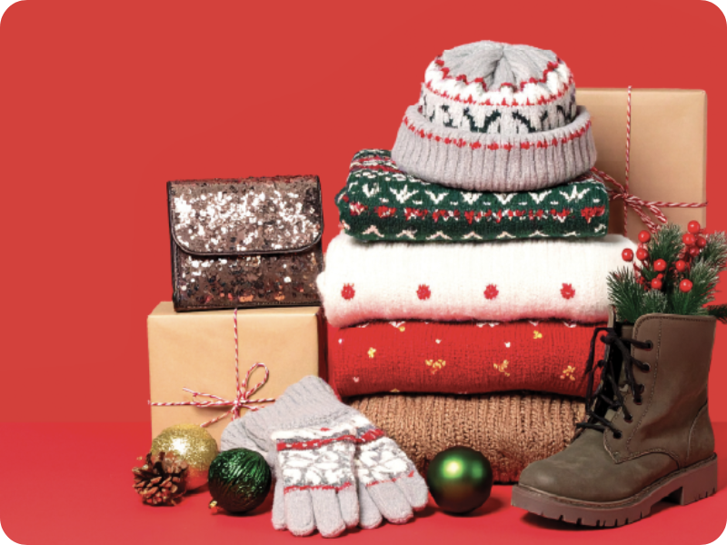 Stack of festive knitwear, gloves and gifts with Christmas ornaments, a sequinned clutch bag and winter boots against a red background.