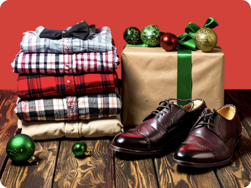 Folded plaid shirts, wrapped gifts, festive ornaments and polished dress shoes arranged on a wooden floor against a red background.