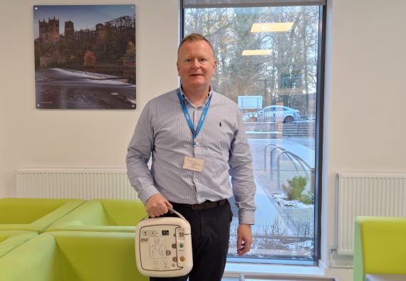 A man stood smiling and holding a defibrillator case in his right hand
