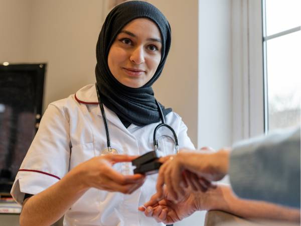 A smiling nurse wearing a headscarf uses a blood pressure monitor to check a patient’s arm during a health check.