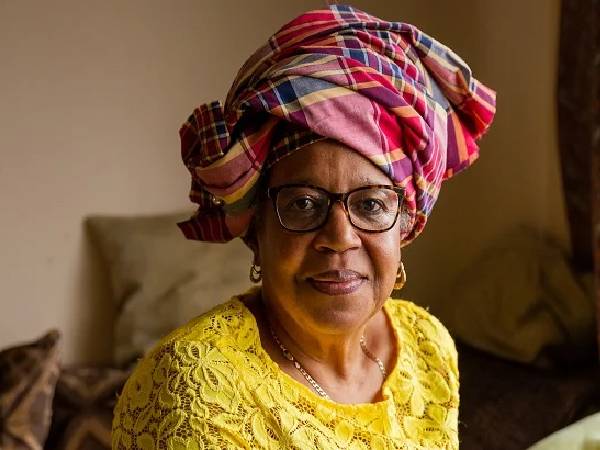 A close up image of an older black woman sitting in her living room . She is wearing a bright yellow dress and a red, patterned headwap and is smiling contentedly at the camera.