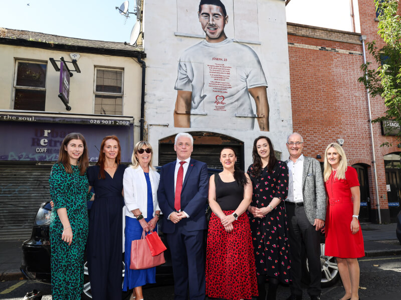 A group of people stand outside, and a large mural can be seen behind them. 