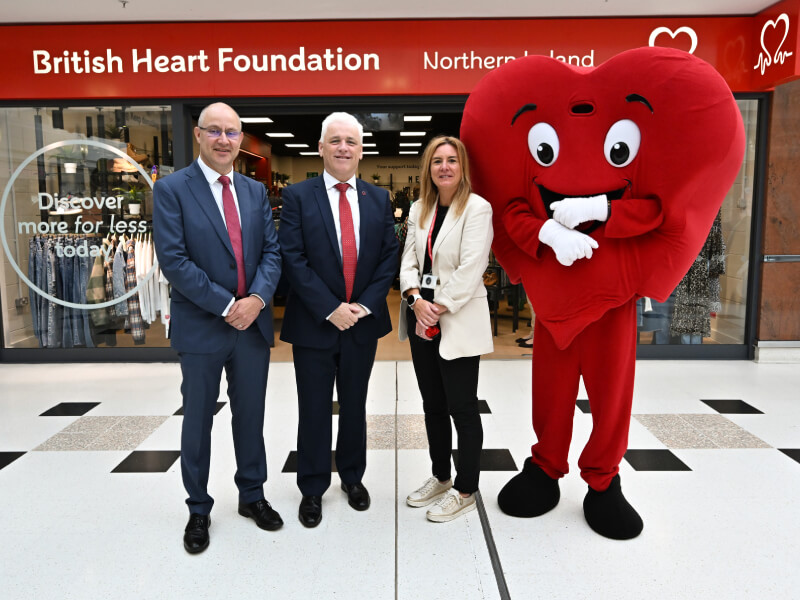 Three people stand outside a British Heart Foundation shop in Northern Ireland, with a mascot dressed as a giant red heart