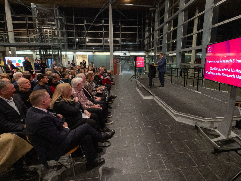 A male conference speaker stands at a podium and presents to an audience sat in front of him. 