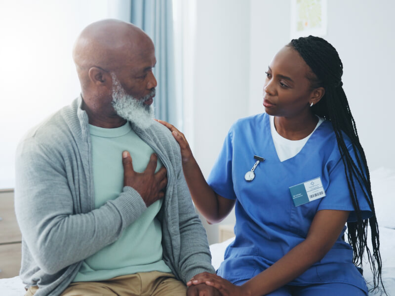 A female nurse in blue scrubs comforts a male patient who is clutching his heart