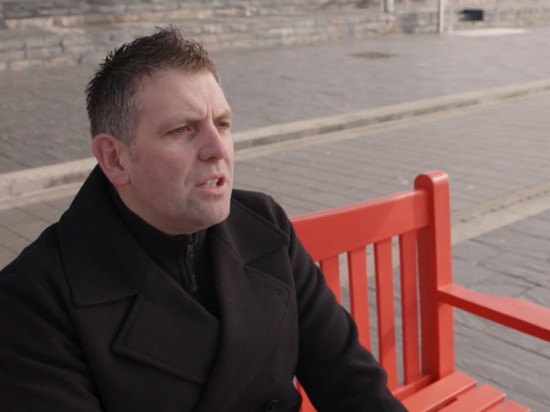 Dyn yn eistedd ar fainc goch yn sgwrsio â rhywun