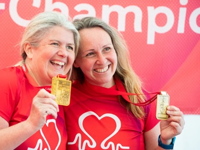 Dwy fenyw mewn crysau T BHF, yn gwenu ac yn dangos medalau aur