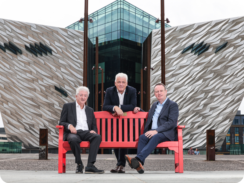 Manufacturing NI CEO Stephen Kelly and Chair of Enterprise NI, John McGrillen on a red BHF bench in front of Titanic Belfast, a modern building with a striking geometric silver facade and glass entrance.