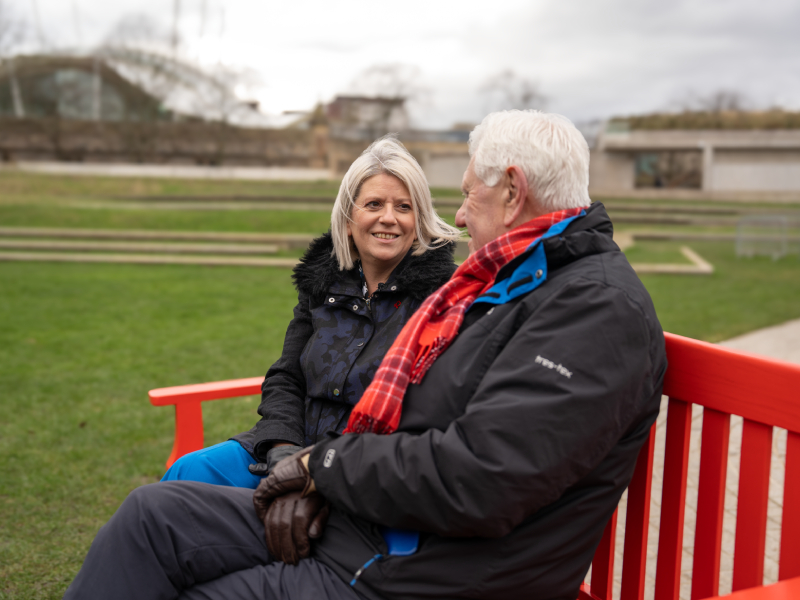 A man and woman sat on a red bench looking at each other and smiling