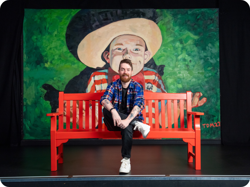 Rosco McClelland sitting on a bright red bench in front of a large painted portrait with a green background, at The Stand Comedy Club in Glasgow.