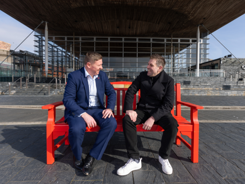 Two men sat on a red bench having a conversation and smiling