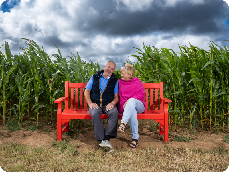 Hugh Thomas sits next to his wife on a red BHF bench beside a tall green cornfield under a cloudy sky.