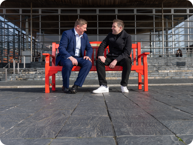 Two people sit on a red BHF bench in front of the Senedd, Cardiff.