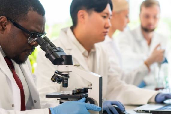a group of researchers wearing white coats in a lab one of whom is looking through a microscope