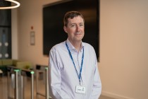 Photograph of BHF Professor Declan O'Regan. He is looking into the camera. Behind him we can see out of focus entry gates. In the photo is wearing a shirt with an open collar and a lanyard with his ID card.