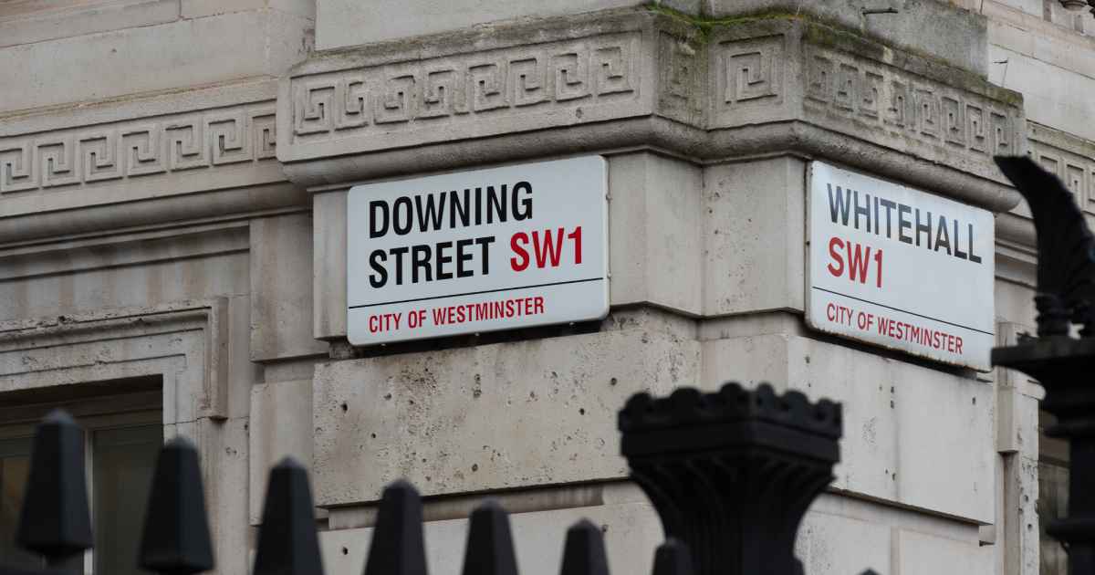 Street signs on a stone building corner read ‘Downing Street SW1’ and ‘Whitehall SW1’ in the City of Westminster, with a black metal fence in the foreground.