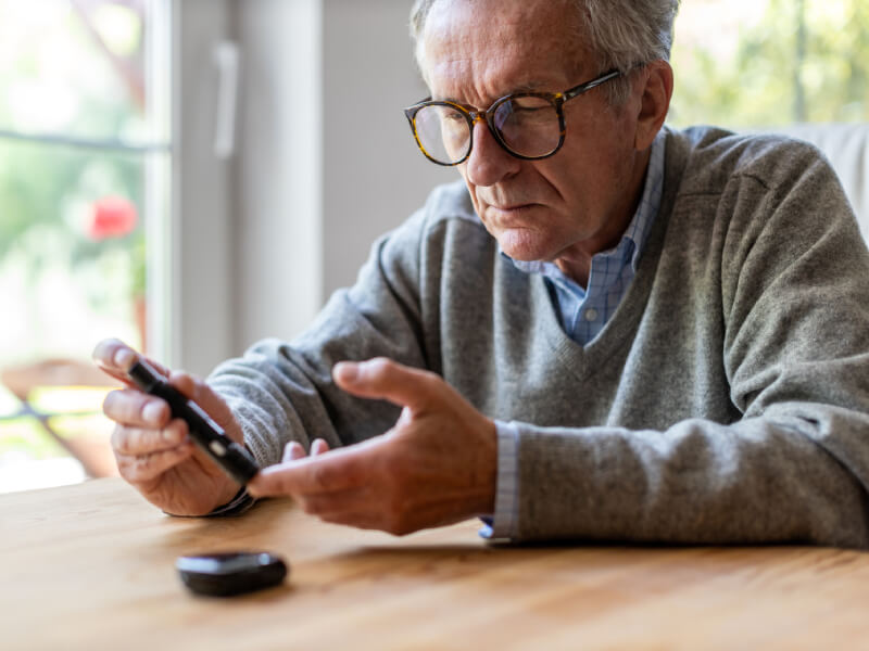 An old man sits at a table and takes a blood sugar reading