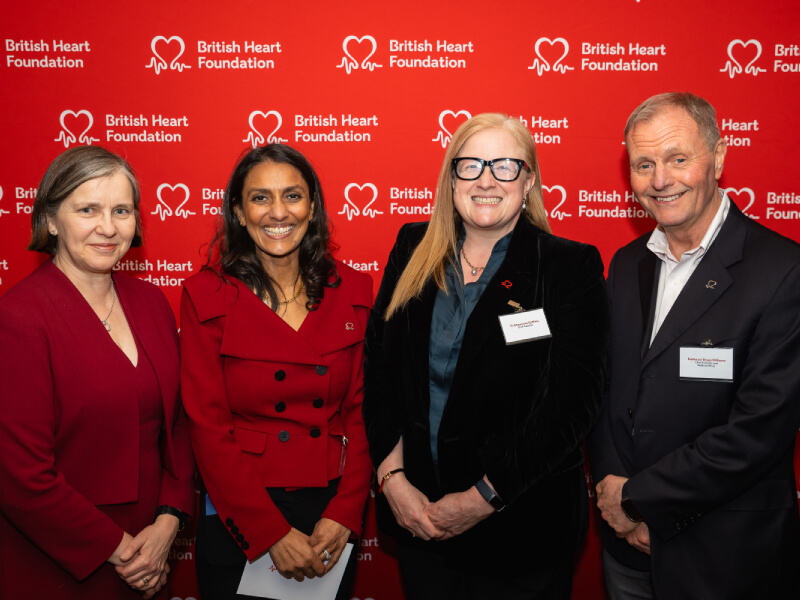 4 people stand and pose for a photograph in front of a red backdrop featuring the BHF logo