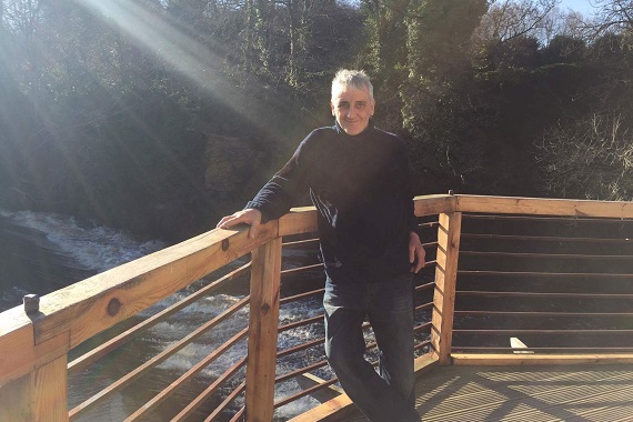 Steve Hawkins stands outside on a wooden terrace by a river.