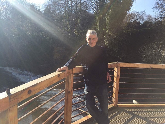Steve Hawkins stands outside on a wooden terrace by a river.