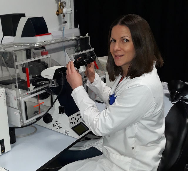 Dr Sarah Jones, researcher at Manchester Metropolitan University sits at a desk using a microscope. She is wearing a lab coat and looking at the camera.