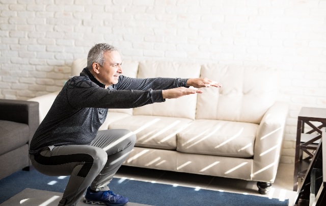 A man squatting in a living room on an exercise mat with his arms raised and smiling.