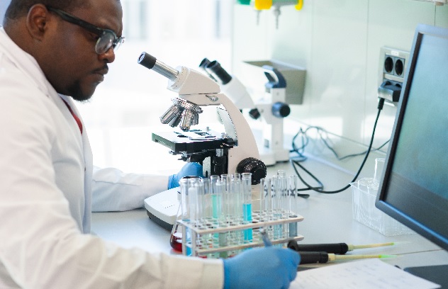 A man sits in front of a microscope with a set of viles holding liquids while writing notes.