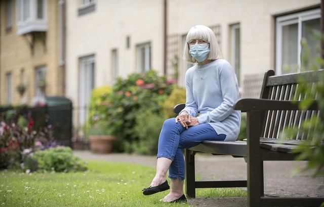 A woman with grey hair in her 60s sitting on a dark brown bench in a garden