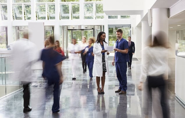 A busy hospital where everyone is blurred except a female doctor talking to a male nurse in the entrance to a hospital