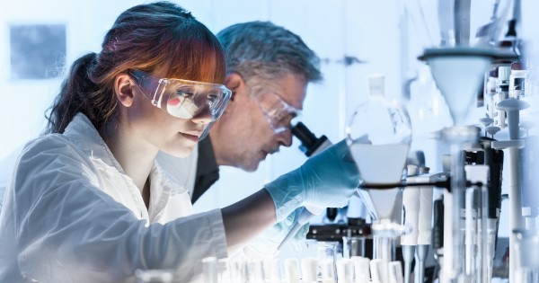 Two people in white coats in a laboratory surrounded by test tubes, one is looking down a microscope and the other is using a pipette