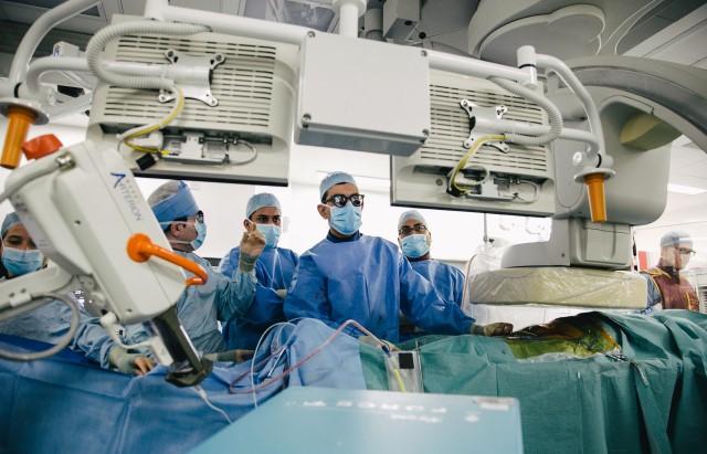 A group of surgeons stand beside the bed of a patient during an operation looking at a screen