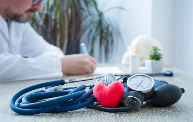 A blood pressure monitor on a table with a red heart toy resting on it. A healthcare professional writes at a desk in the background.