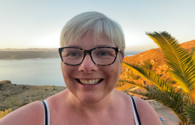 A woman takes a selfie at sunset with a palm tree and the sea behind her.