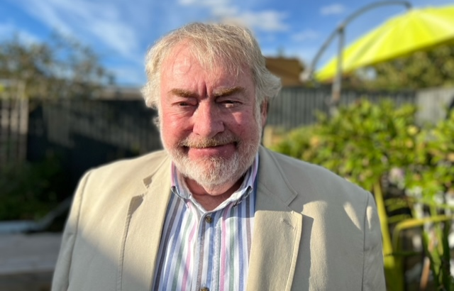 Headshot of a man in a cream suit in a sunny back garden.
