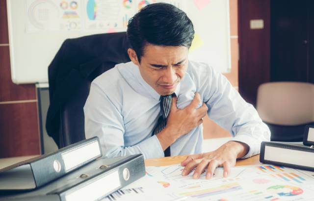 A man in a shirt and tie behind a desk clutches his chest in pain.