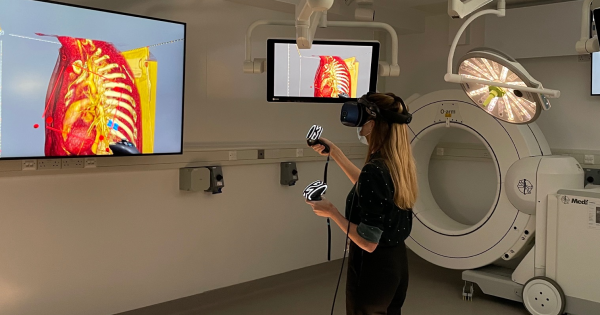 A researcher wears a Virtual Reality headset with a controller in each hand. They're viewing a 3D recreation of a patients heart, which is projected onto screens around them.