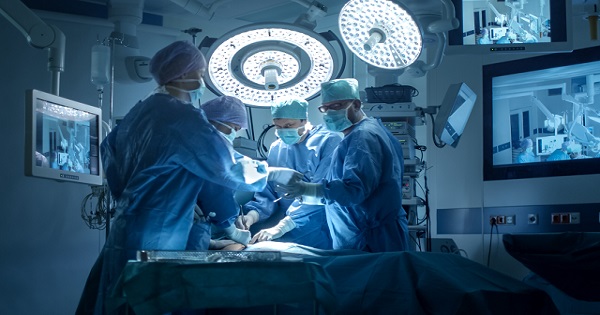 Four surgeons wearing blue scrubs, face masks, and caps, backlit by bright lights surrounded by hopsital monitors in an operating theatre