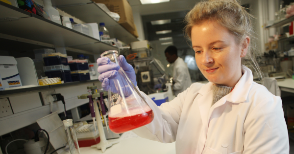 Dr Karla O'Neill stands in a lab holding a beaker containing red liquid