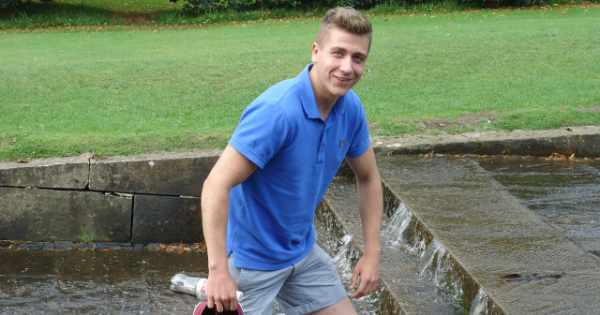 Photograph of Stephen Hughes wearing a blue tshirt and grey shorts carrying a pair of red trainers as he walks through a water feature at Chatworth House