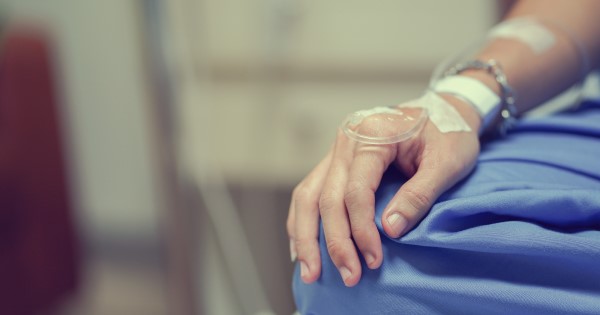 Cropped image of a patient in a blue hospital gown sits with their hand on their knee and a drip in the back of their hand