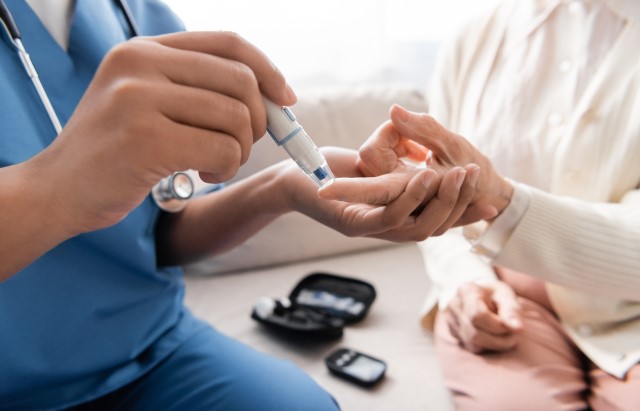 We see a medical worker wearing a blue scrub and a stethoscope around his neck, and a patient wearing a white blouse, cardigan, and pink pants. They are sitting next to each other on the sofa. The medical professional holds the device on the tip of the patient's left index finger and pricks the skin to draw a drop of blood. This drop of blood is then used to test the patient's blood sugar levels.