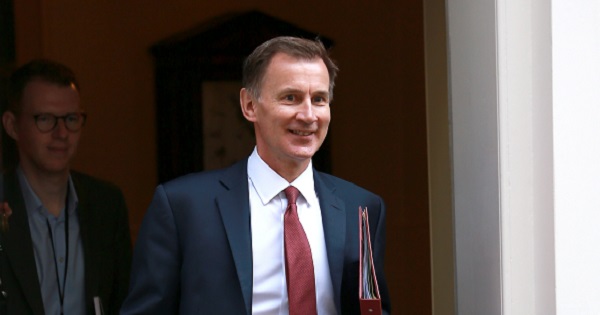 A close cropped image of Chancellor Jeremy Hunt leaving a building through an open door. He is a white male in his late 50s, has short brown hair and is wearing a navy suit with a white shirt and red tie, He is carrying a red folder under his left arm. Someone is leaving the building behind him.