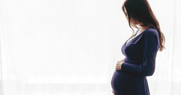 A pregnant woman wearing a blue dress stands in front of a white drape holding her belly