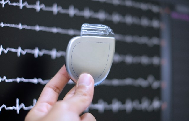 A photo of an ICD, which someone is holding between their thumb and index finger. The ICD is silver. It is in front of a monitor showing the trace of an ECG, a test which measures a person's heart rhythm.