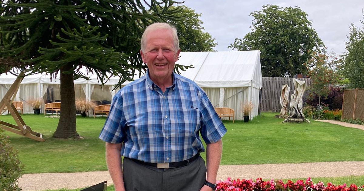 A photograph of Peter Dyde, a participant in a BHF clinical trial Peter is standing in a garden smiling at the camera. He is wearing a blue checked shirt with grey trousers. Behind him we can see a tree, a marquee and benches.